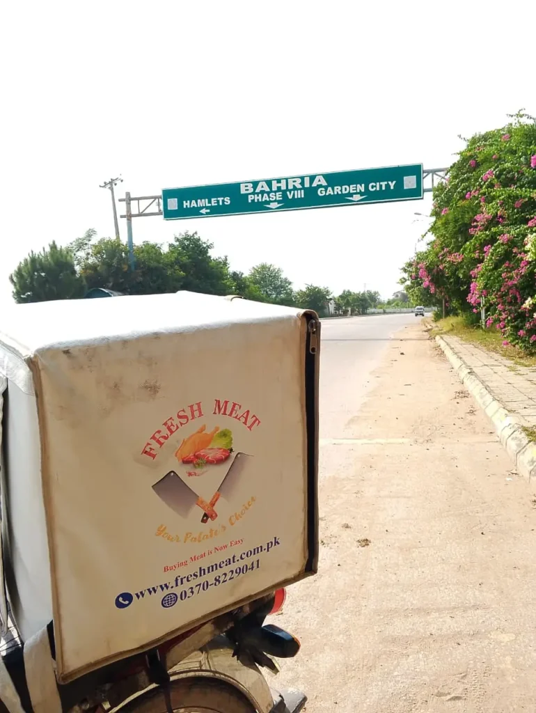 Bahria road sign pointing to Garden City, Phase VIII and Hamlets, with a Fresh Meat delivery bike